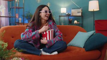 Young woman sits on couch eating popcorn, watching interesting TV serial sport game online at home video