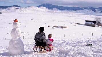 Mother with a small child sits on a sled near a snowman. Back view video