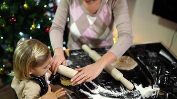 Mom rolls out cookie cutters with a rolling pin on the dough on the table next to a little girl video