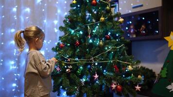 Little girl hangs a garland on the branches of a Christmas tree over toys and balls video