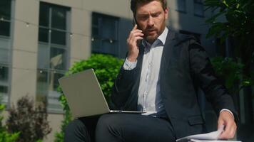 Multitasking busy Caucasian middle-aged businessman business man male guy entrepreneur in city talking mobile phone call using laptop working job paperwork document papers checking discuss startup video