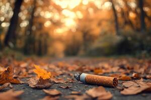 close-up of unextinguished cigarette lies on ground with autumn leaves in forest road with blurred background at evening photo