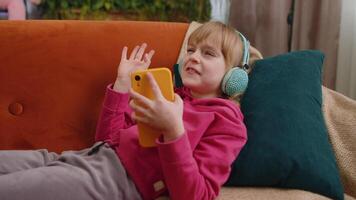 Relaxed child kid girl in headphones listening energetic disco music in smartphone lying on sofa video