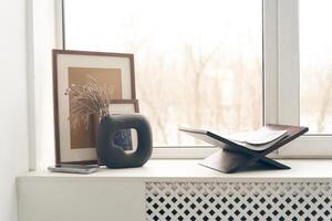 Minimalist composition on the windowsill near the window with a copy space. A vase with dried flowers, a book, and photo frames. Home Decor Template