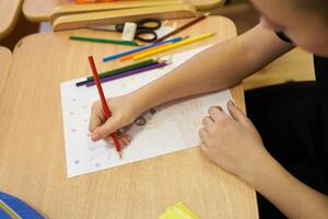 On a white sheet of paper, an unrecognizable child is intently drawing at a table with a set of colored pencils. With space to copy photo