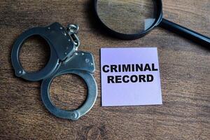 Concept of Criminal Record write on sticky notes and handcuff isolated on Wooden Table. photo