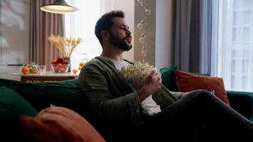 Caucasian bearded joyful man business male businessman guy homeowner buyer client customer falling on sofa couch relaxing eating popcorn unhealthy snack watching TV television using controller at home video