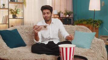 Indian man on sofa eating popcorn, watching movie on smartphone focused on screen enjoying free time video