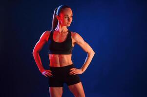 Hands are on the waist, standing. Beautiful muscular woman is indoors in the studio with neon lighting photo