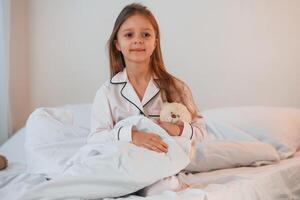 Little, girl, with teddy bear in hands is sitting on the bed at home photo
