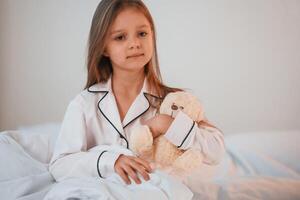 Little girl with teddy bear in hands is sitting on the bed at home photo