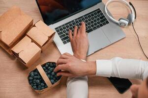 Taking blueberries. Top view of hands, of the man that is working by using laptop. Eco boxes with food photo