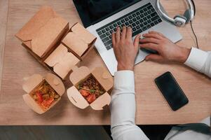 Top view of man's hands that is, typing on the, laptop. Food in the paper eco boxes are on the table photo