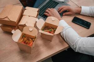 Top view of man's hands tha,t is typing on the laptop. Food in the paper eco boxes are on the table photo