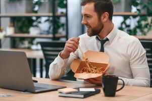 Sitting by laptop. Businessman,, in formal clothes is in the office eating udon that is in the eco box photo