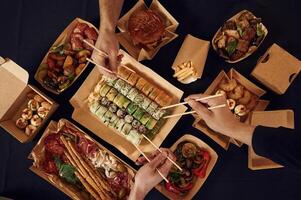 Top view of table, with food ,on it, hands of the people taking it, eating. Meat, sushi, vegetables, French fries, burger, mushrooms photo