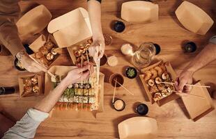 Top view of table, with sushi on it in paper eco boxes, hands of the people taking it, eating photo