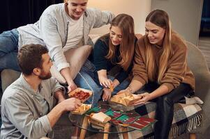 On ,the sofa.. Man is sitting on the floor. Friends are sitting indoors at home and eating together photo