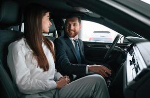 Man, in suit and tie is sitting with young woman, customer, in the automobile and showing the interior of the car photo