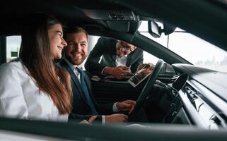 Side, view. Car showroom worker is consulting man and woman that are sitting inside of automobile photo