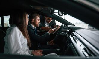 Side view. Car showroom, worker, is consulting man and woman that are sitting inside of automobile photo