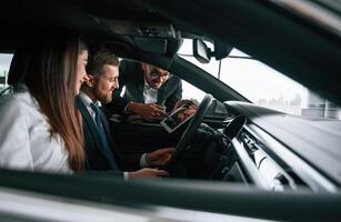 Side view. Car showroom worker is ,consulting man and woman that are sitting inside of automobile photo