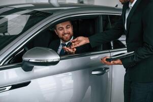 Salesman gives keys of new automobile to the customer, that is smiling and sitting in the car photo