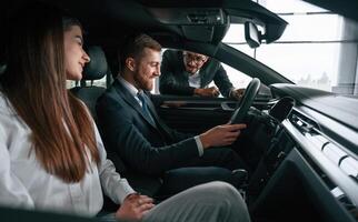 Side view.. Car showroom worker is consulting man and woman that are sitting inside of automobile photo