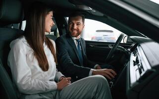 Man in, suit and tie is sitting with young woman, customer, in the automobile and showing the interior of the car photo