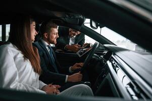 Car showroom ,worker is consulting man and woman that are sitting inside of automobile photo