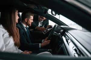 Car ,showroom worker is consulting man and woman that are sitting inside of automobile photo