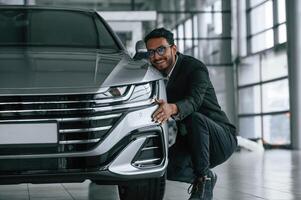 Embracing ,the transport. Handsome indian man in suit is in the car showroom with the vehicle indoors photo