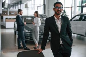 Man standing, in front of his coworkers. Three people are working together in the car showroom photo