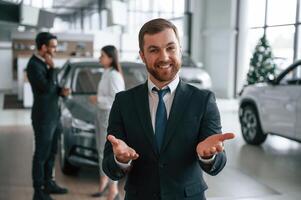 Beautiful man is standing, and smiling. Three, people are working together in the car showroom photo