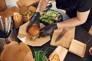 Close up view, of burger, French fries and vegetables. Man is packing food into the paper eco boxes. Indoors, restaurant photo