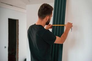 Taking, measurements.. Plumber worker installing heating radiator in empty room of a newly built house photo