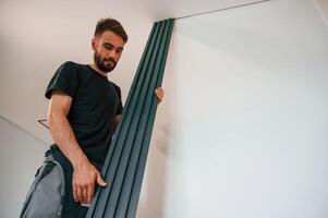 Holding tall heating radiator. Plumber worker is in empty room of a newly built house photo