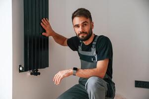 Looking at, the ,camera. Plumber worker installing heating radiator in empty room of a newly built house photo