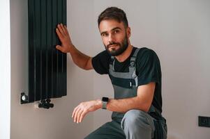 Looking at the camera. Plumber worker installing heating radiator in empty room of a newly built house photo