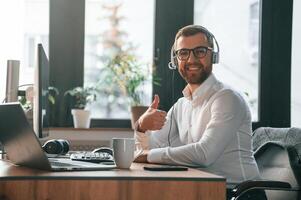 Smiling, sitting by table. In headphones. Office worker in formal clothes is sitting by computer and working photo