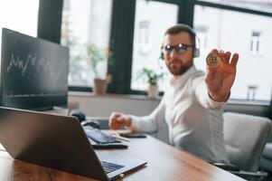 Sitting and showing physical bitcoin. Man in formal clothes is working in the modern office. Using computer photo