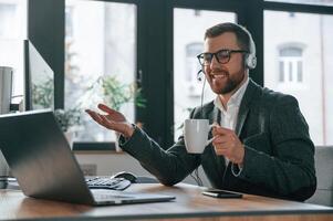 In headphones. Having online conversation. Businessman in suit is in the office with computer photo