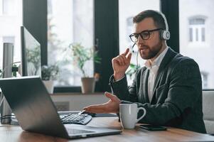 In headphones.. Having online conversation. Businessman in suit is in the office with computer photo