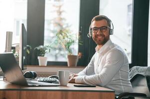 Smiling,, sitting by table. In headphones. Office worker in formal clothes is sitting by computer and working photo