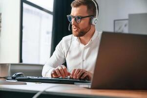 In, headphones. Man, in formal clothes is working in the modern office. Using computer photo