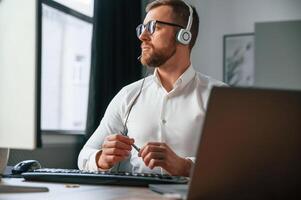 In headphones.. Man in formal clothes is working in the modern office. Using computer photo