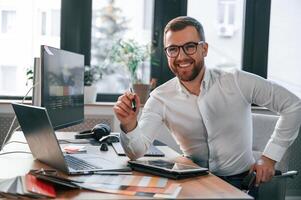 Sitting and, smiling. Man in formal clothes is working in the modern office. Using computer photo