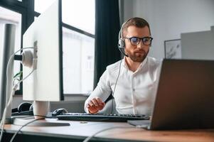 In headphones. Man, in formal clothes is working in the modern office. Using computer photo