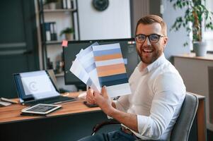 Designer is choosing different colors for project. Man in formal clothes is working in the modern office. Using computer photo