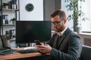 Sitting, using smartphone. Businessman in suit is in the office with computer photo